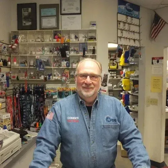 Man in denim shirt smiles in a store. Shelves with merchandise are in the background.