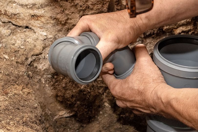 Hands fitting gray PVC pipes together in a dirt trench during underground plumbing work