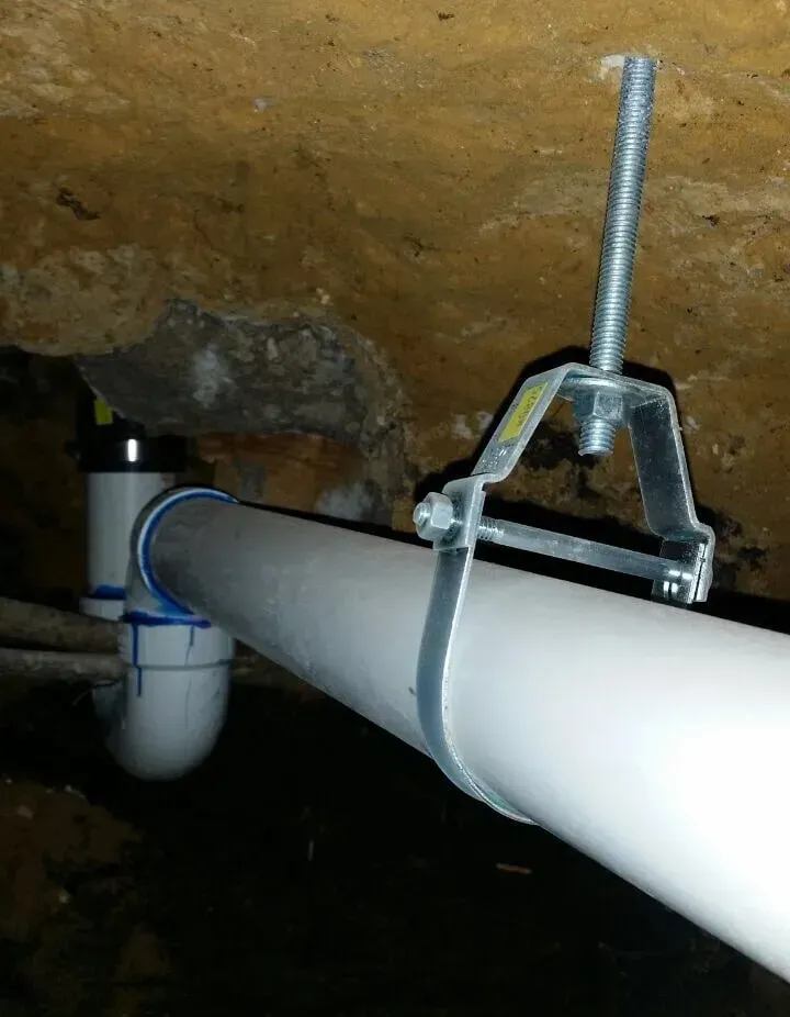 White pipe clamped to a rocky ceiling with a metal bracket in a dark utility space