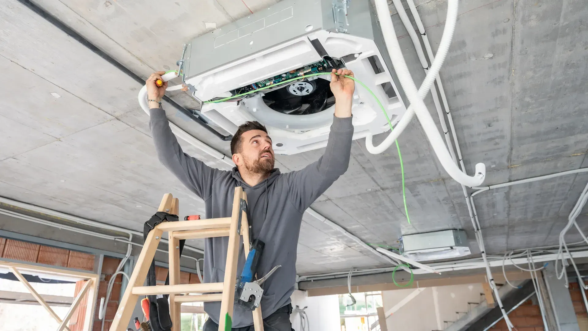 Man in gray shirt installing a ceiling-mounted unit on a ladder in an unfinished room