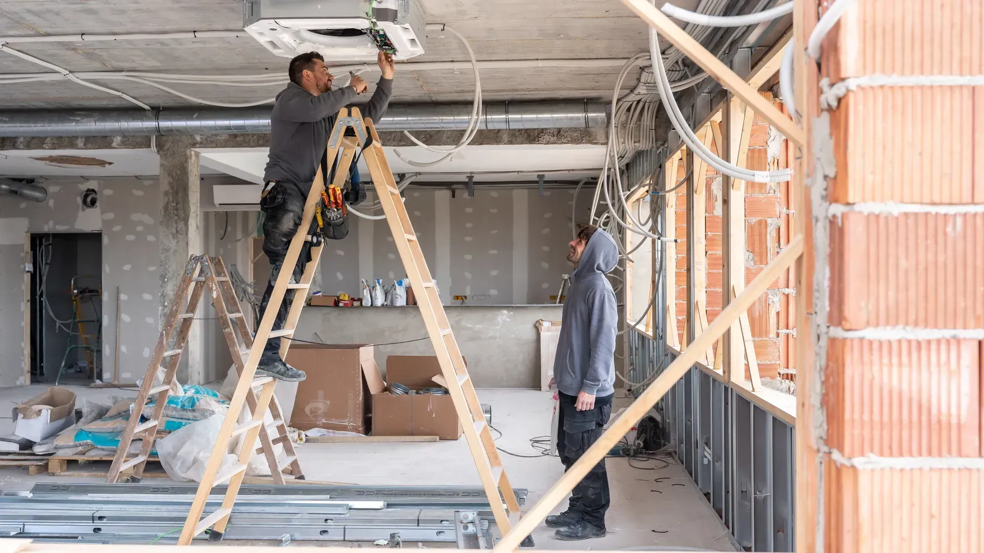 Two workers installing ceiling fixtures in a house under construction, with ladders and exposed framing.