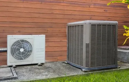 Two outdoor HVAC units on a concrete pad beside a wooden wall and grass