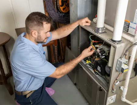Technician kneeling and inspecting an open furnace control panel with tools in a utility room