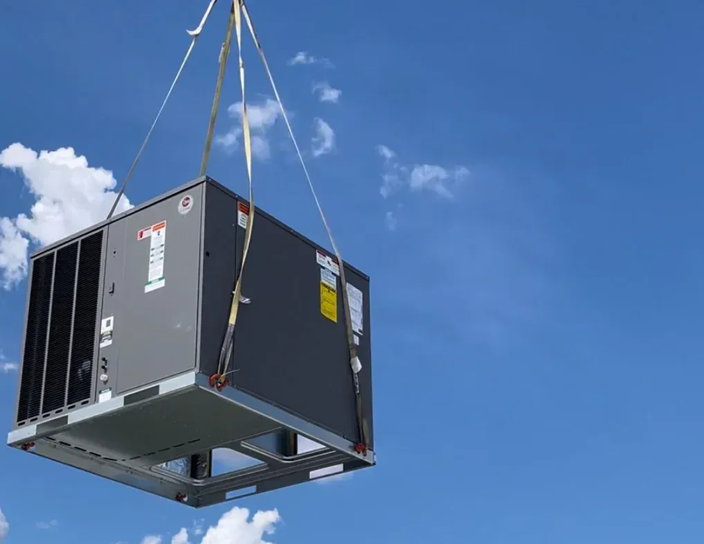 A large black shipping container hangs from straps against a bright blue sky with clouds.