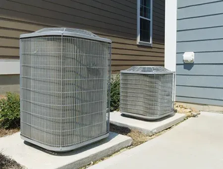 Two outdoor air conditioning units beside a house wall and shrubbery