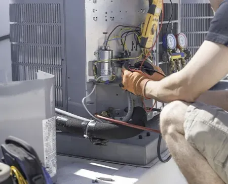 Technician kneels beside HVAC equipment, working with tools and gauges on an outdoor unit.