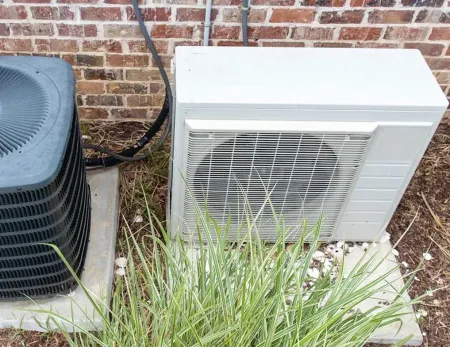 Outdoor HVAC units beside a brick wall, with tall grass in the foreground