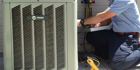 Technician kneels beside HVAC unit, working with tools and wires indoors.