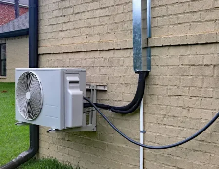 Wall-mounted outdoor HVAC unit with black cables on a beige brick building exterior.