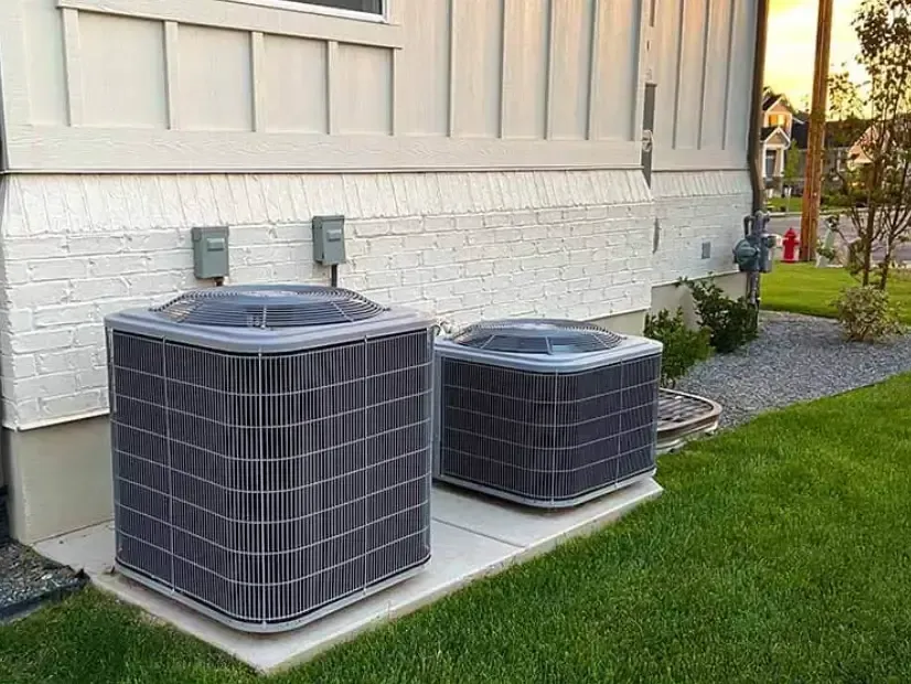 Two gray outdoor AC units beside a white house on a concrete pad, next to green lawn.