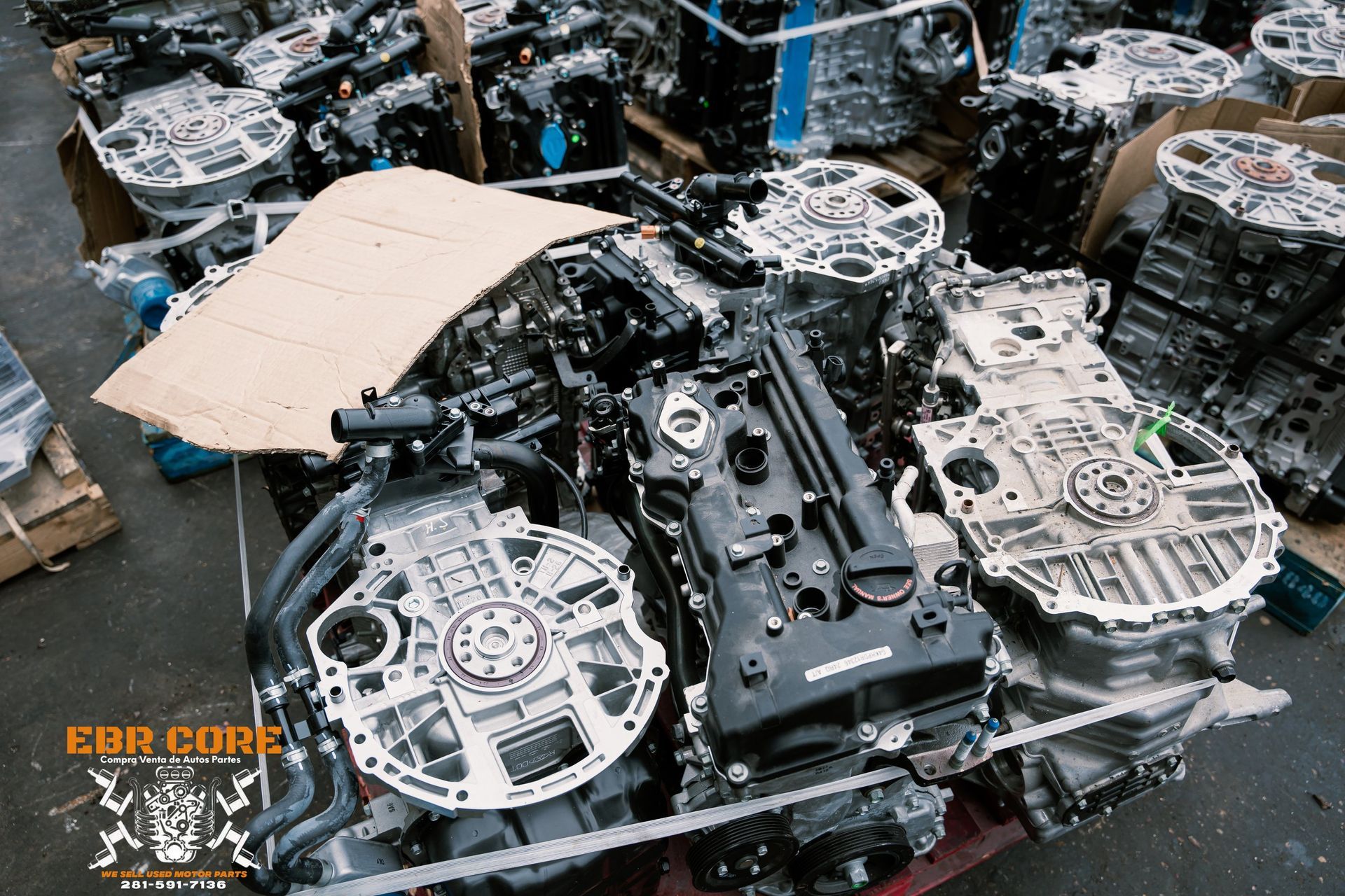 Pile of used car engines. Mostly black and gray metal parts, cardboard, inside a warehouse.