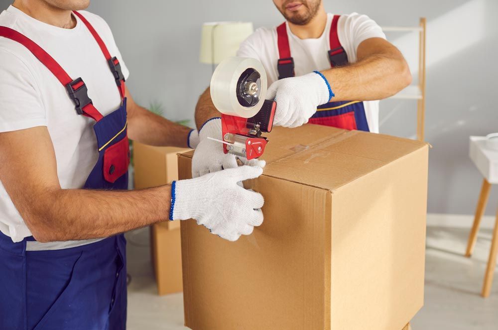 Two Movers in Blue Overalls Sealing a Cardboard Box — Greater Newcastle Removals In Rathmines, NSW