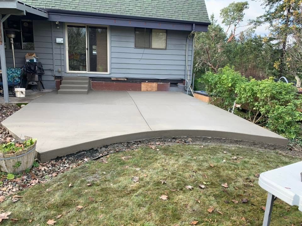 Concrete patio in front of a gray house, with a curved edge and garden beds nearby