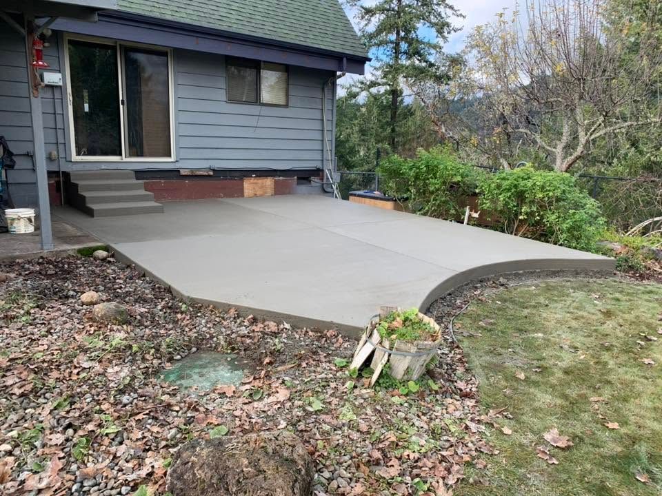 Concrete patio beside gray house with curved edge, bordering lawn and shrubs.