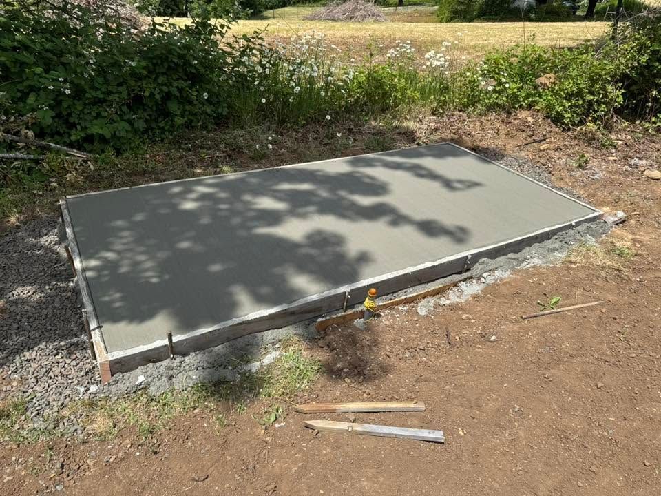Concrete slab foundation in a dirt yard with trees and shrubs nearby, partly in shadow.