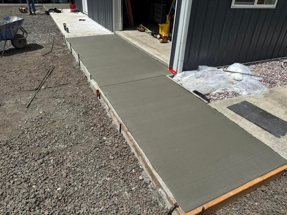 Fresh concrete walkway being finished beside a gray building, with gravel on one side and snow patches nearby