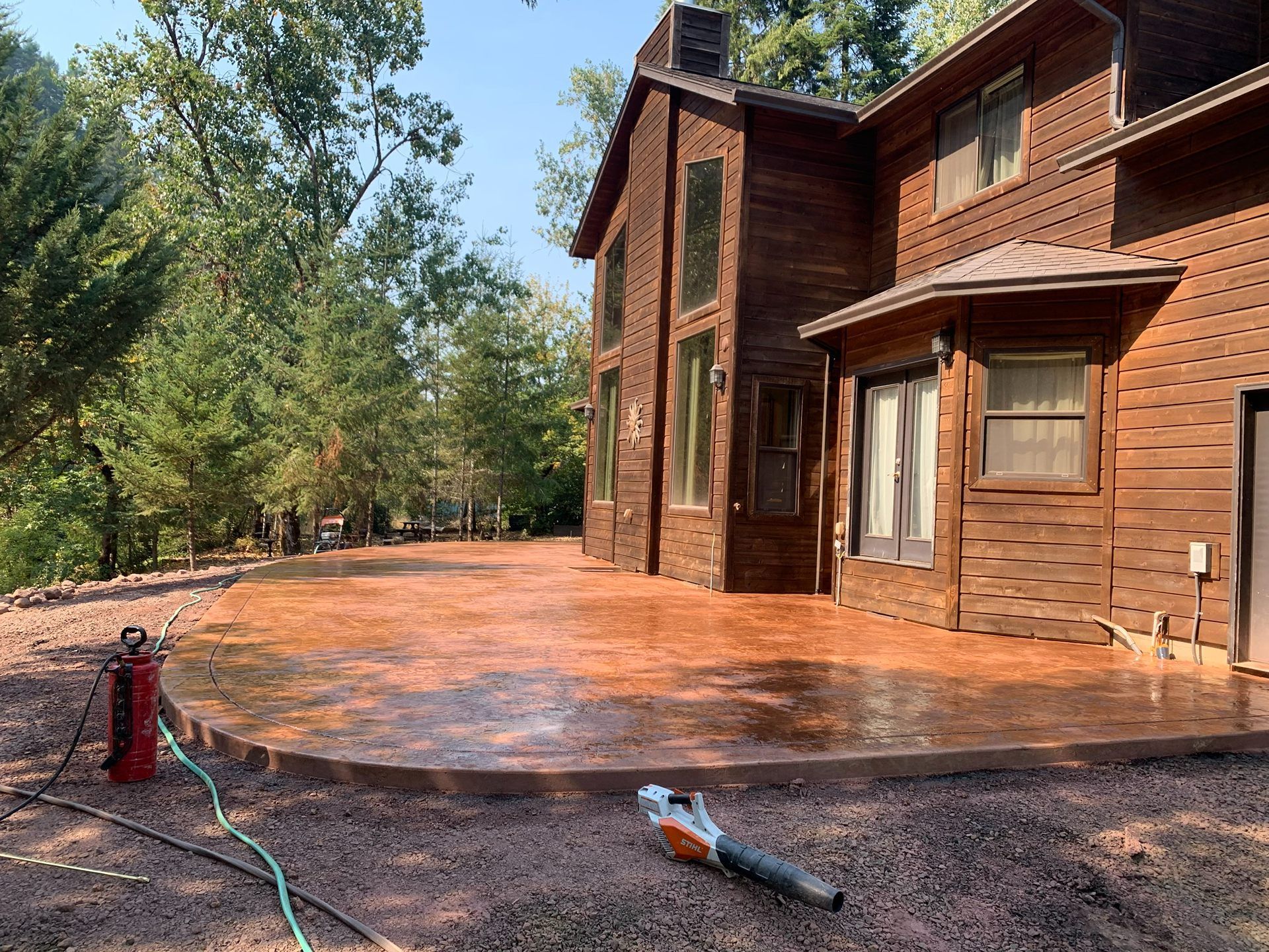 Wooden house with a freshly stained deck, surrounded by trees; tools and a hose lie on the ground.