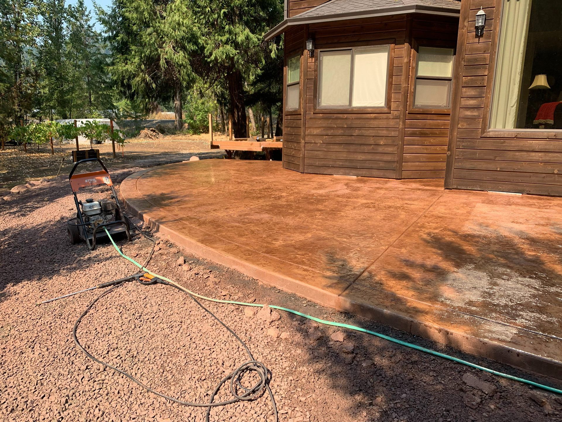 Brown house exterior with a dirt yard, garden hose, and outdoor tools in a wooded area