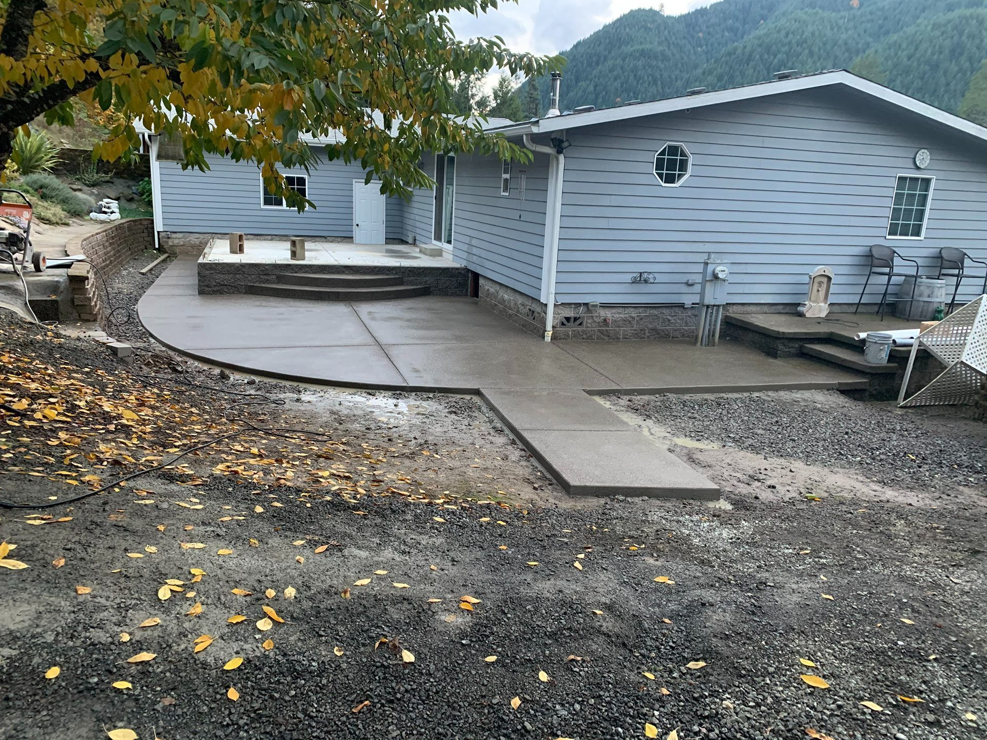 Curved concrete walkway beside a white house with a small deck and gravel yard, under autumn trees