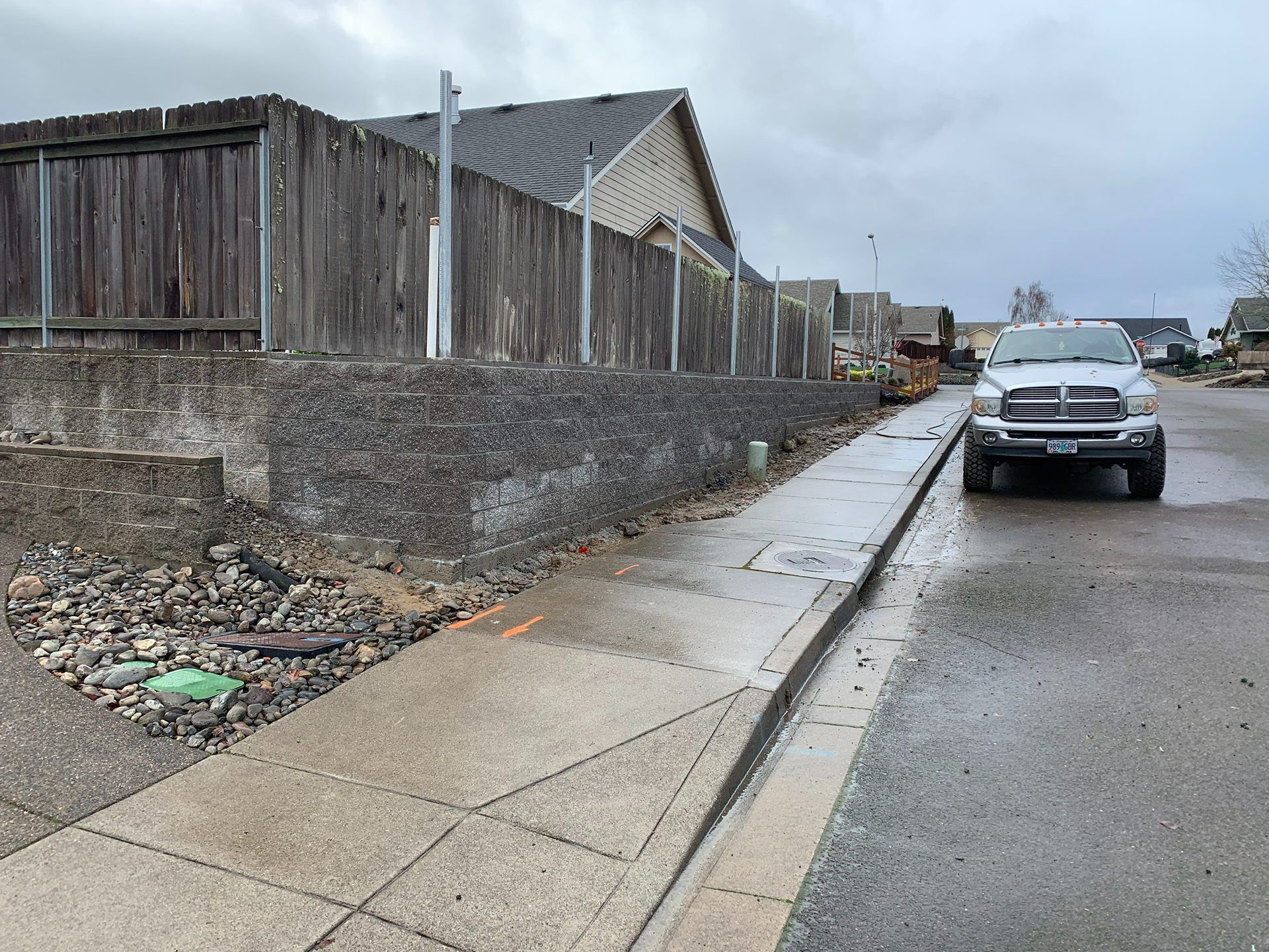 Sidewalk beside a stone wall and wooden fence on a rainy street, with a parked car ahead