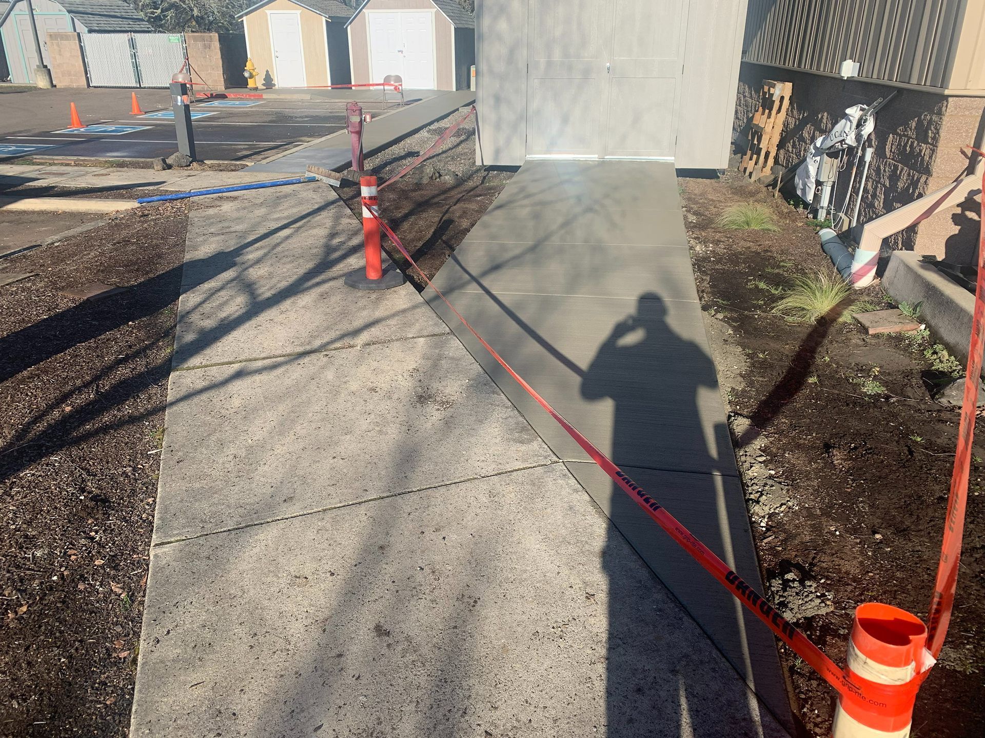 Marked walkway under construction with red caution tape, cones, and a shadow on the pavement