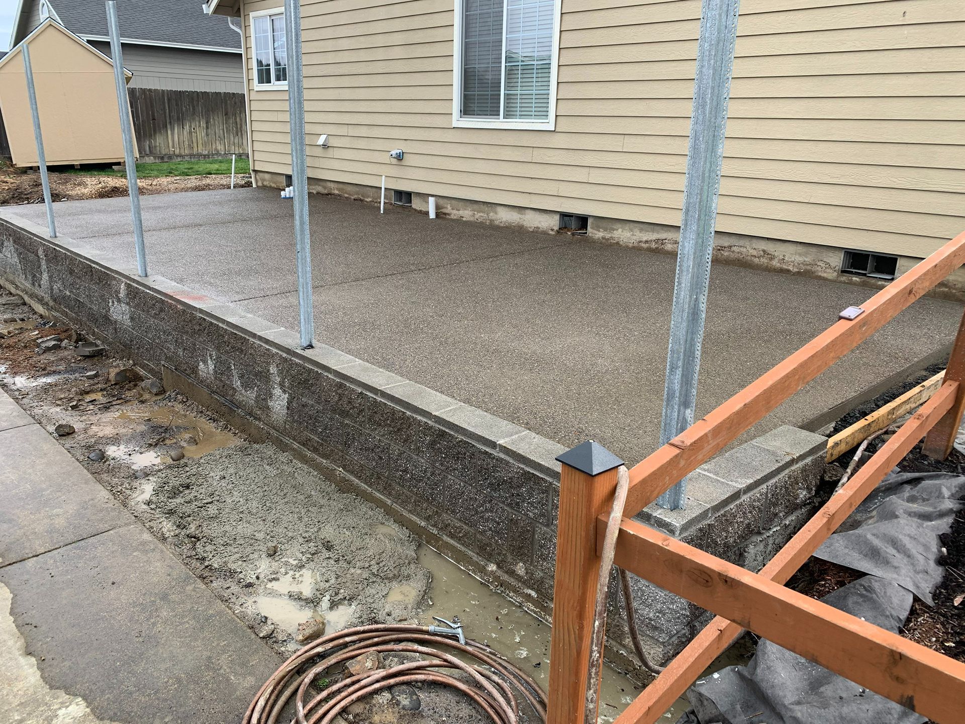 Backyard concrete patio and low retaining wall beside a beige house, with metal railings and wooden framing.