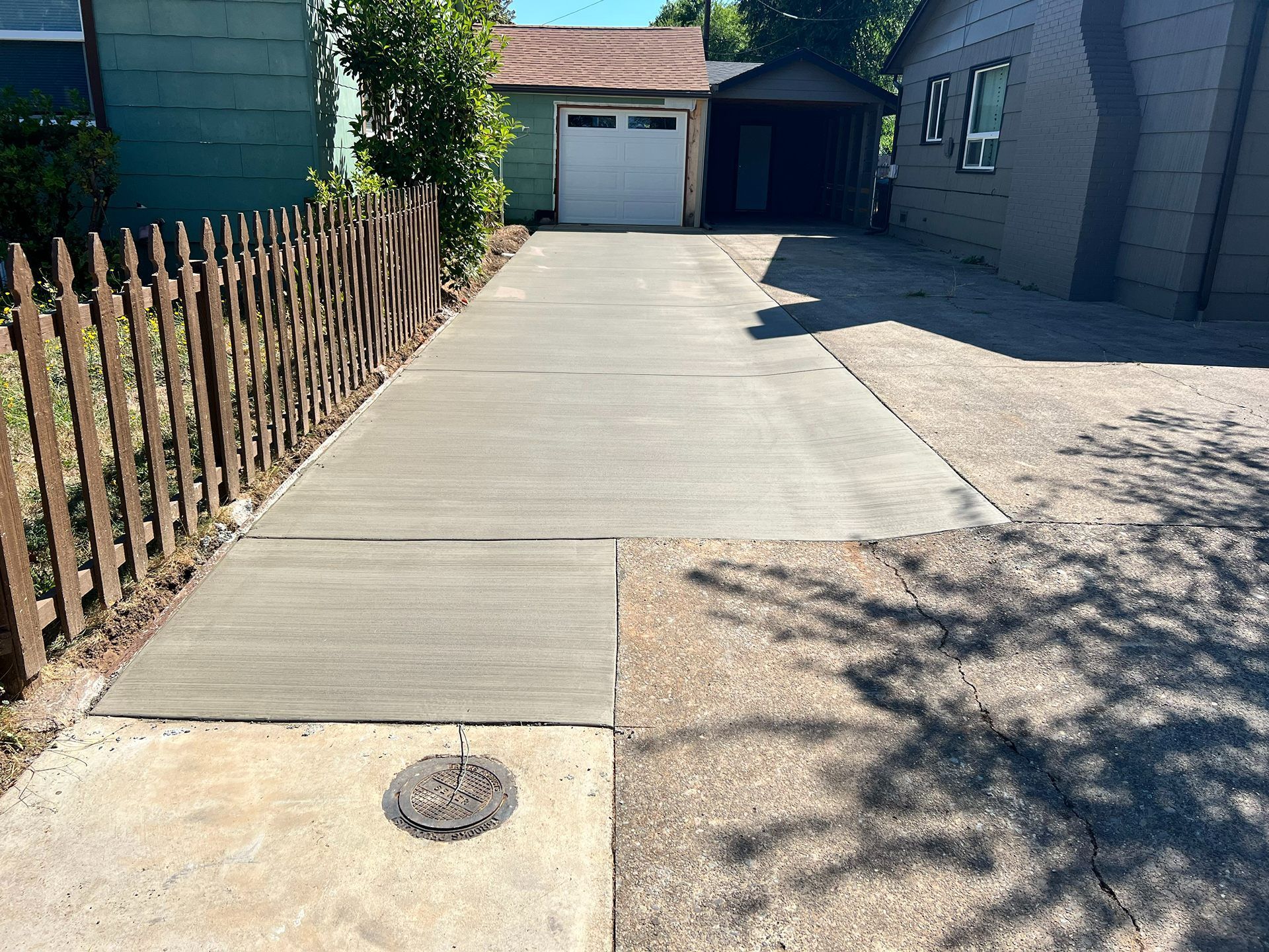 Concrete driveway leading to a garage beside a wooden fence and house, with a drain cover in front.