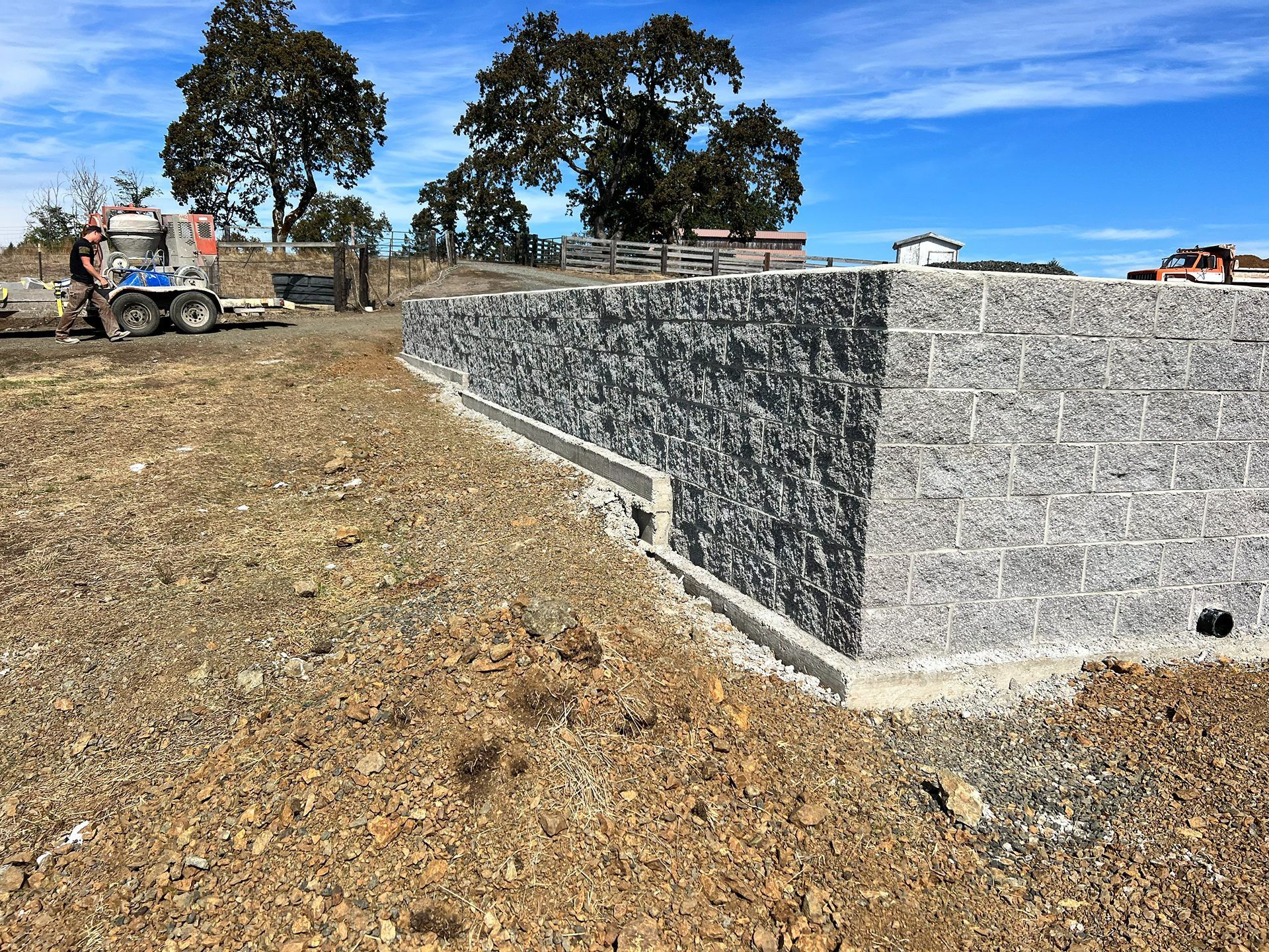 Stone retaining wall under construction on a dry hillside with machinery and trees in the background