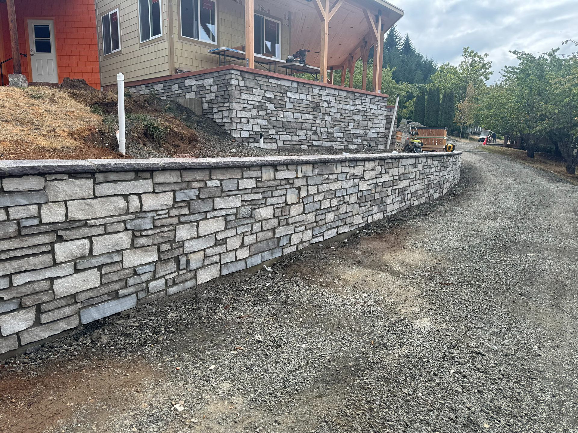 Stone retaining wall beside a gravel driveway under a house with a porch and wooden posts