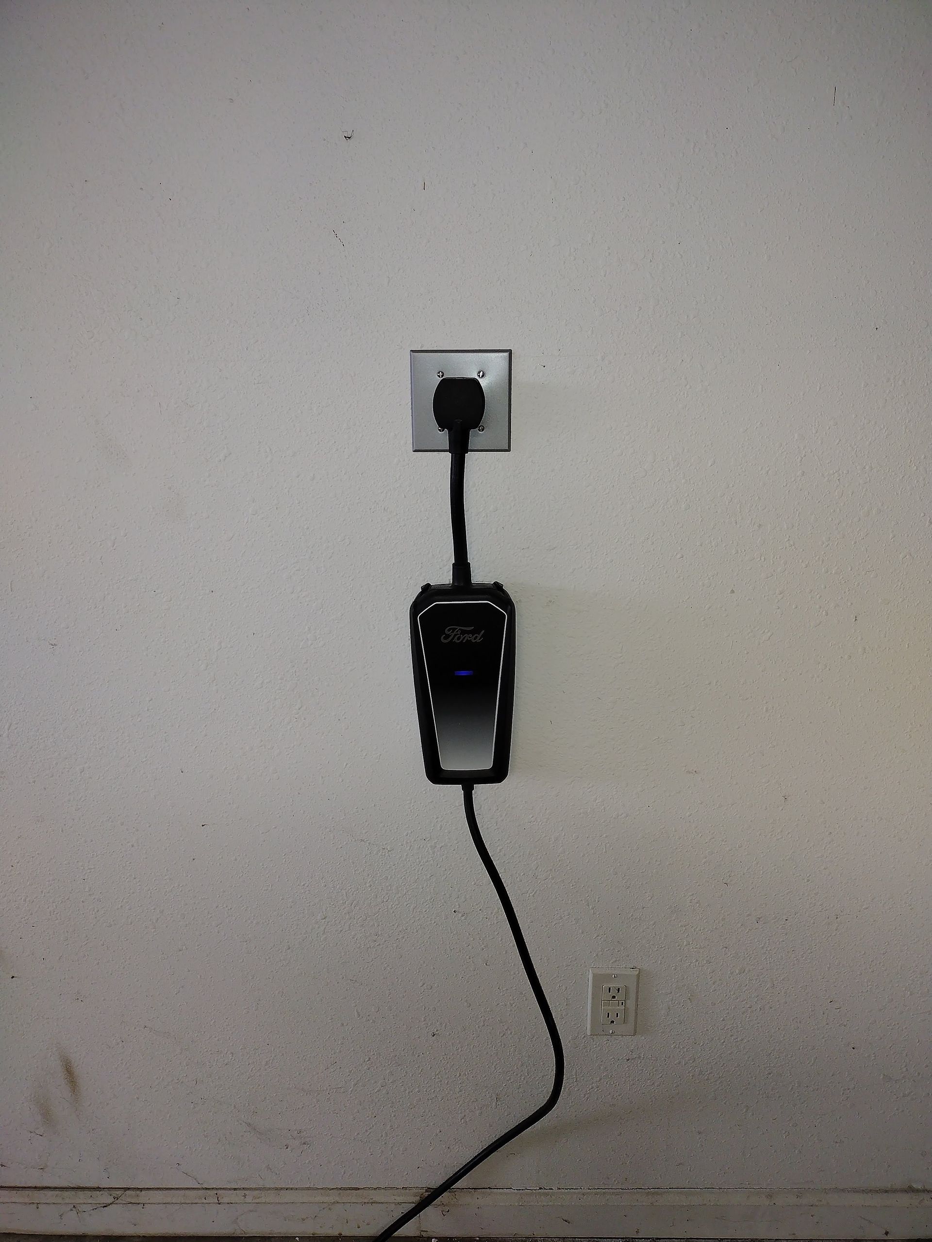Black phone mounted on a wall outlet, with its cord hanging down against a plain wall.