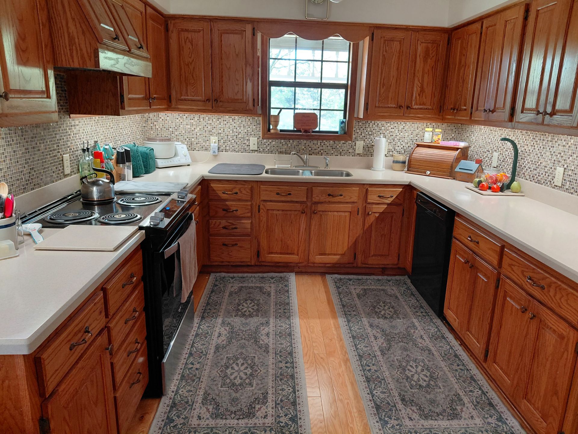 Wooden kitchen with white countertops, patterned backsplash, and a central window above the sink.