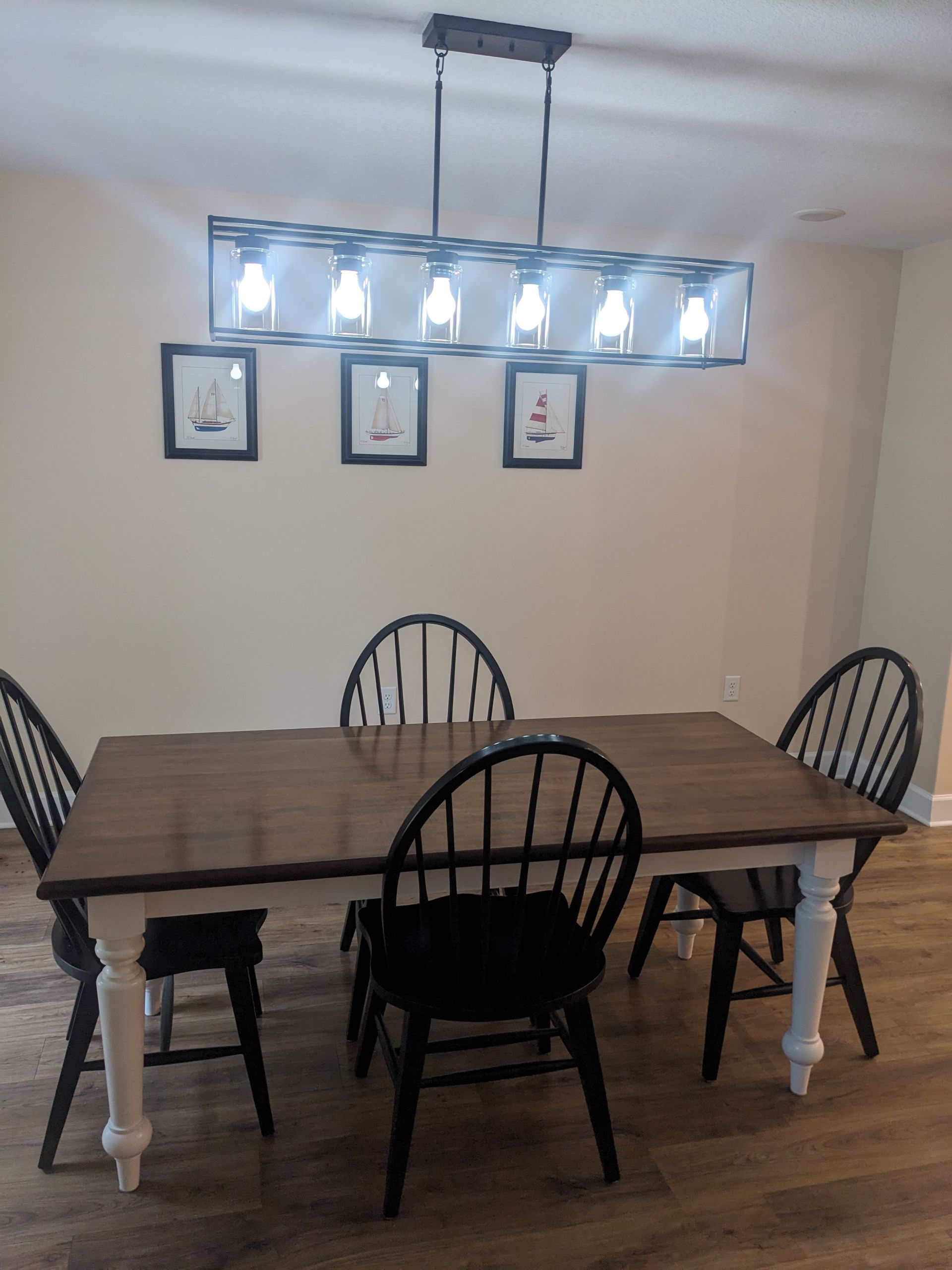 Dining room with a wooden table, four black chairs, framed wall art, and a bright hanging light fixture.