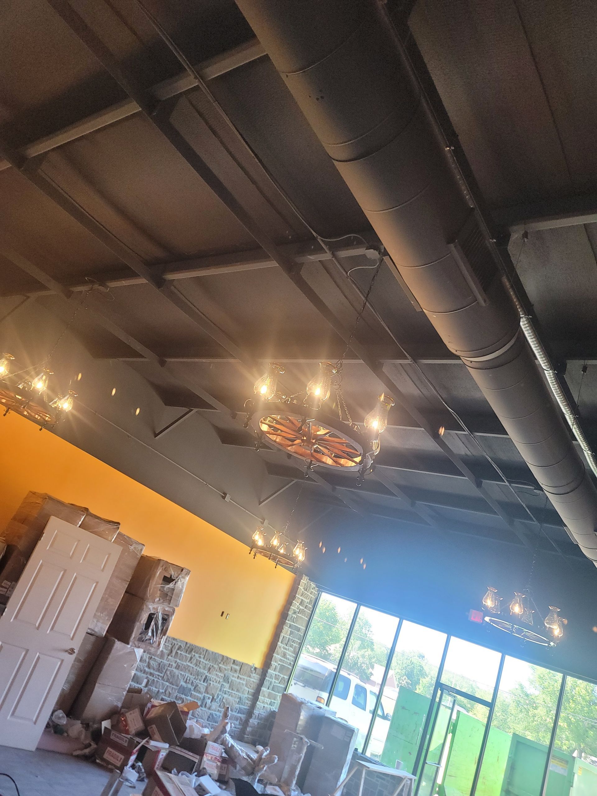 Indoor café with exposed black ceiling, warm lights, orange wall, and large windows showing greenery.