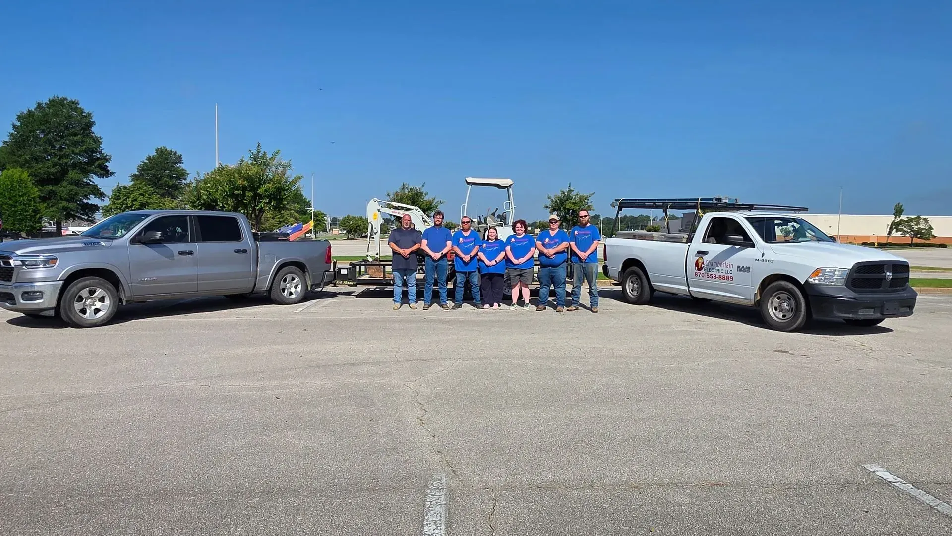 Group of people in matching blue shirts posing between two pickup trucks in a parking lot on a sunny day