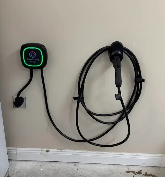 Wall-mounted EV charger with coiled cable and plug on a beige wall above a white baseboard.