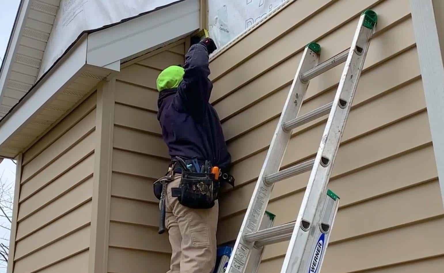 Person on ladder repairing siding on a beige house, wearing a safety vest.