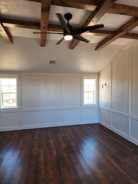Room with dark wood floor, white paneled walls, exposed wooden beams, and a black ceiling fan.
