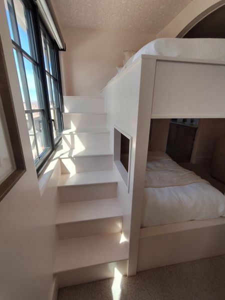 White bunk bed with built-in stairs beside a window, casting shadows.
