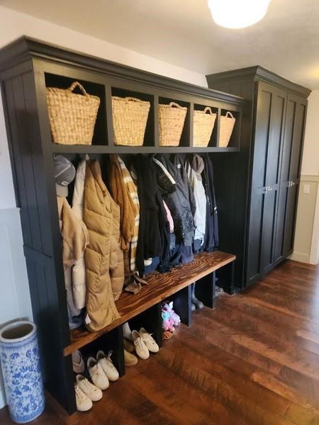 Black entryway storage unit with bench, baskets, and coats. Dark wood floor and tall cabinet.