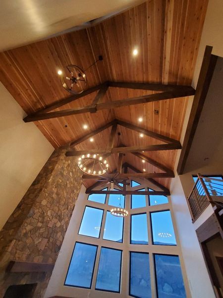 High-ceilinged room with wood beams, cedar ceiling, large windows, and stone fireplace. Three chandeliers hang down.