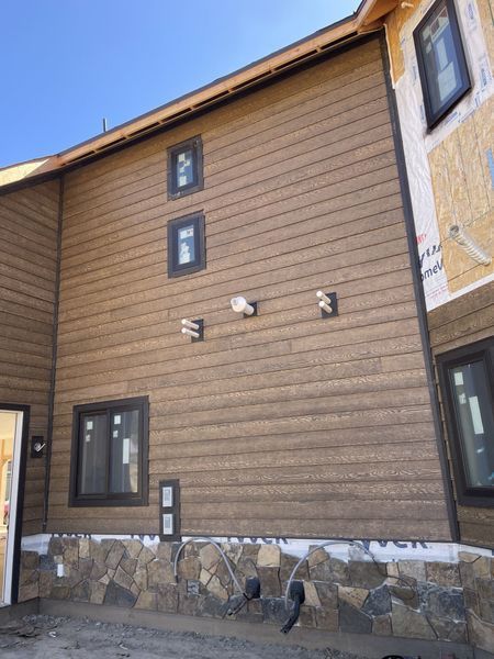 Brown siding on a building under construction with dark trim, windows, and stone foundation.