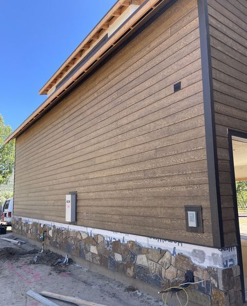Building exterior under construction with brown siding, stone foundation, and dark trim.