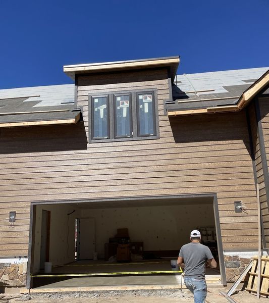 Man walks out of open garage door; new home construction with brown siding, windows, and a blue sky.