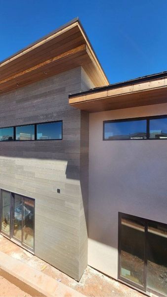 Modern building exterior with gray siding, light wood trim, and large windows against a blue sky.