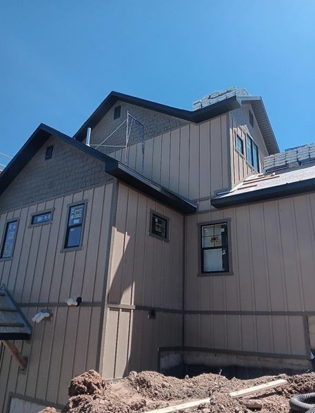Tan and gray two-story house under construction, with dark window frames and a clear blue sky.