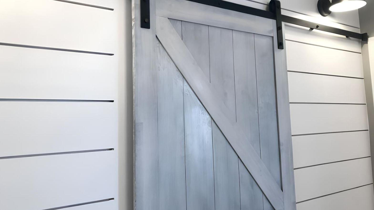 Gray barn-style door with diagonal brace on a white shiplap wall under an exterior light