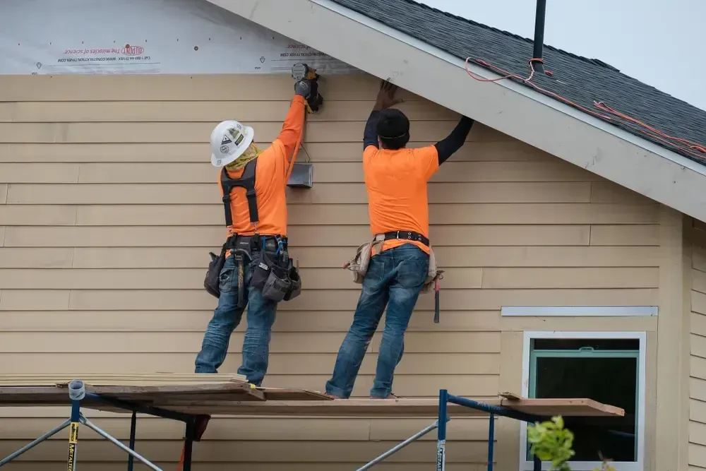 Two construction workers install siding on a house, one using a drill. Orange shirts, blue jeans.