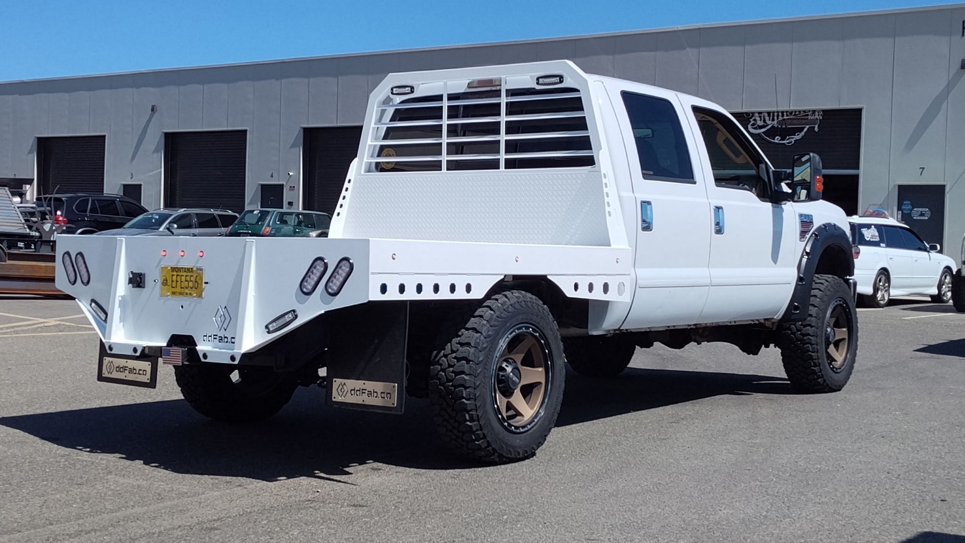 White flatbed truck with large tires parked outside, sunny day.