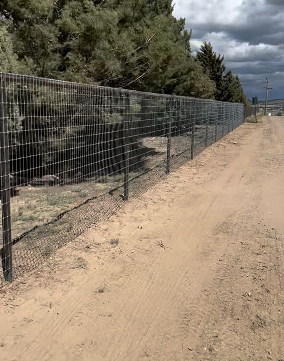 Wire fence alongside a dirt road and greenery, under a cloudy sky.