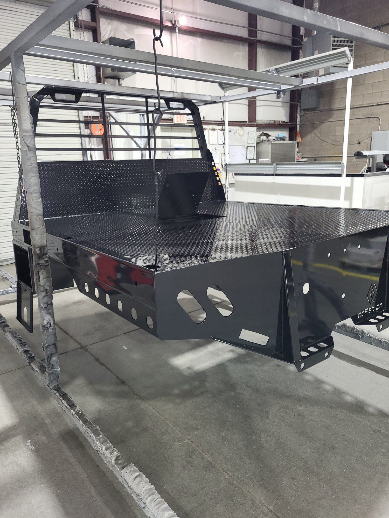 Black metal truck bed on a gray overhead conveyor in an industrial setting.